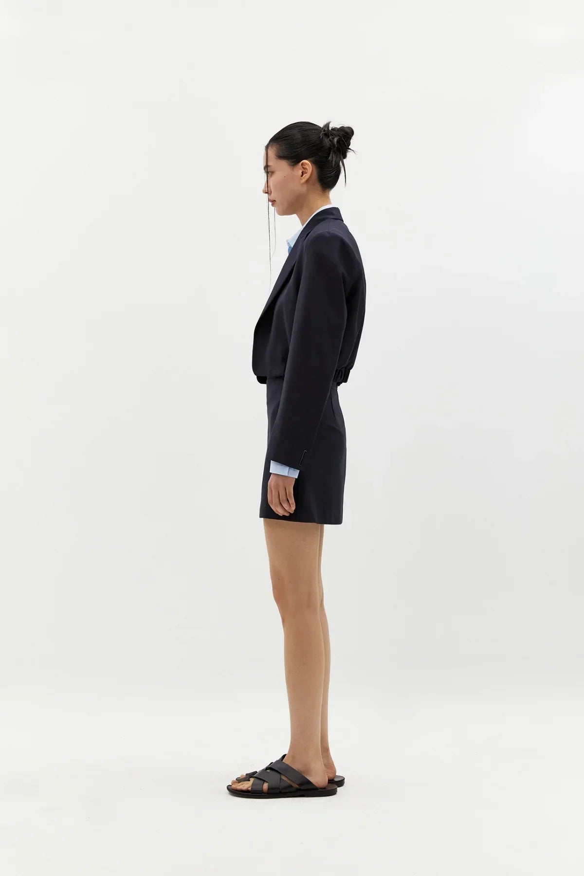 Side view of woman wearing navy cropped blazer and matching short skirt with black sandals on white background