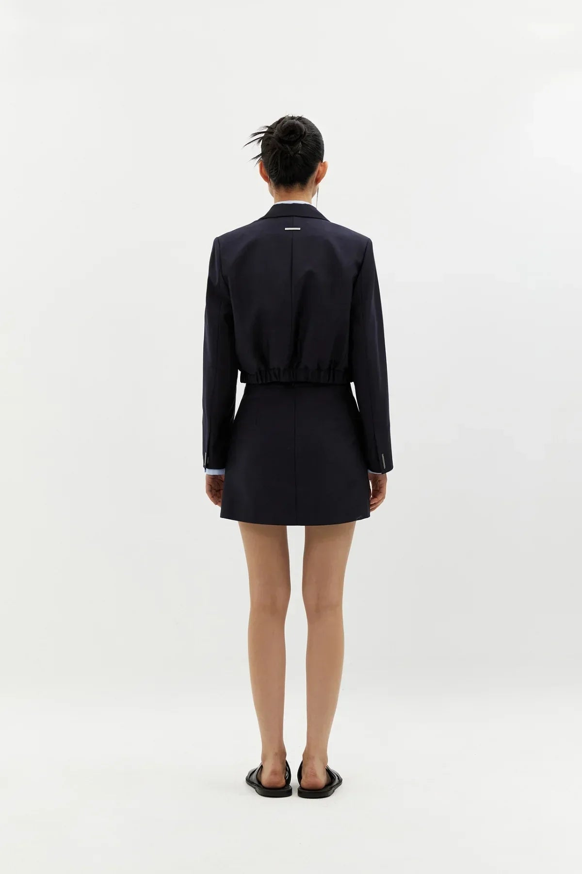 Woman wearing navy short wool skirt and matching cropped blazer with bun hairstyle on white background