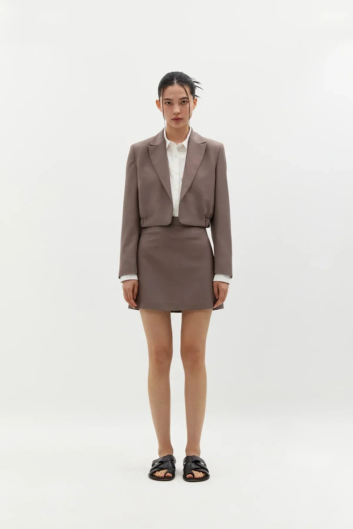 Model wearing taupe cropped blazer and matching short skirt with white shirt and black sandals