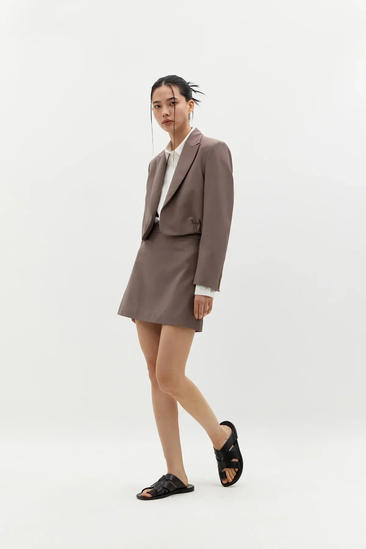 Asian woman in taupe cropped blazer and matching skirt with white shirt and black sandals in studio