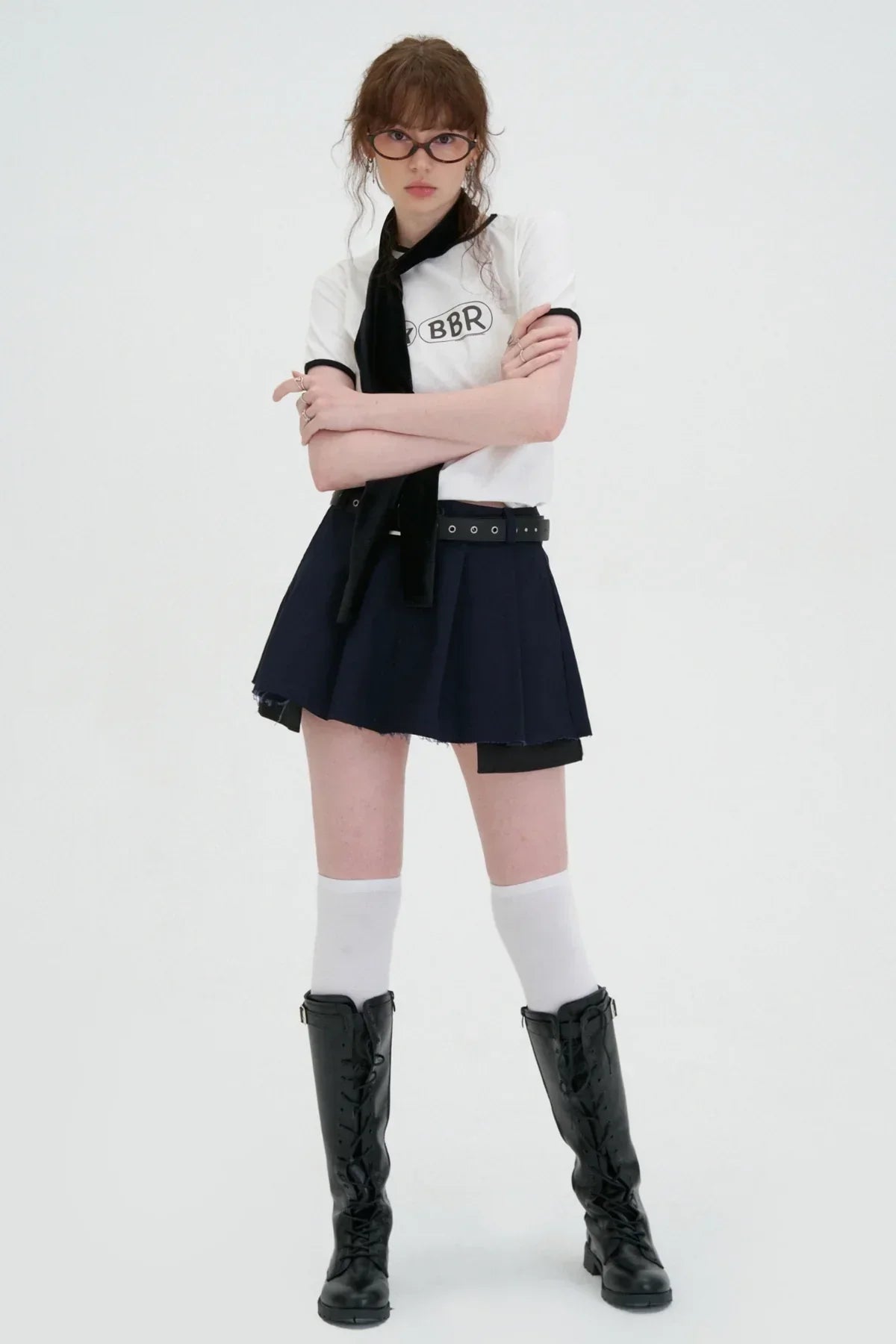 Young woman in white BBR logo t-shirt, navy pleated skirt, knee-high boots, and glasses on white background