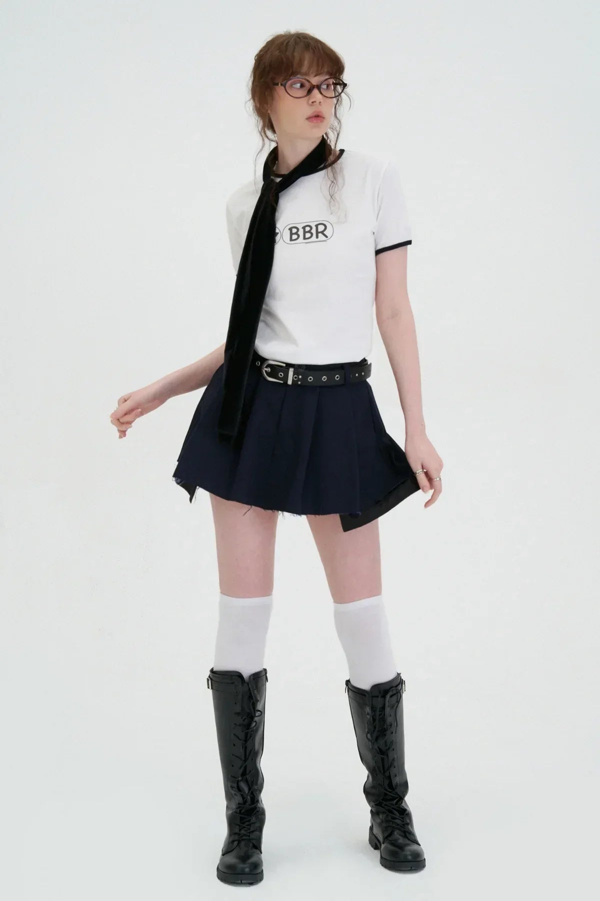 Young woman wearing white BBR logo t-shirt, black pleated skirt, knee-high boots, and white socks in studio