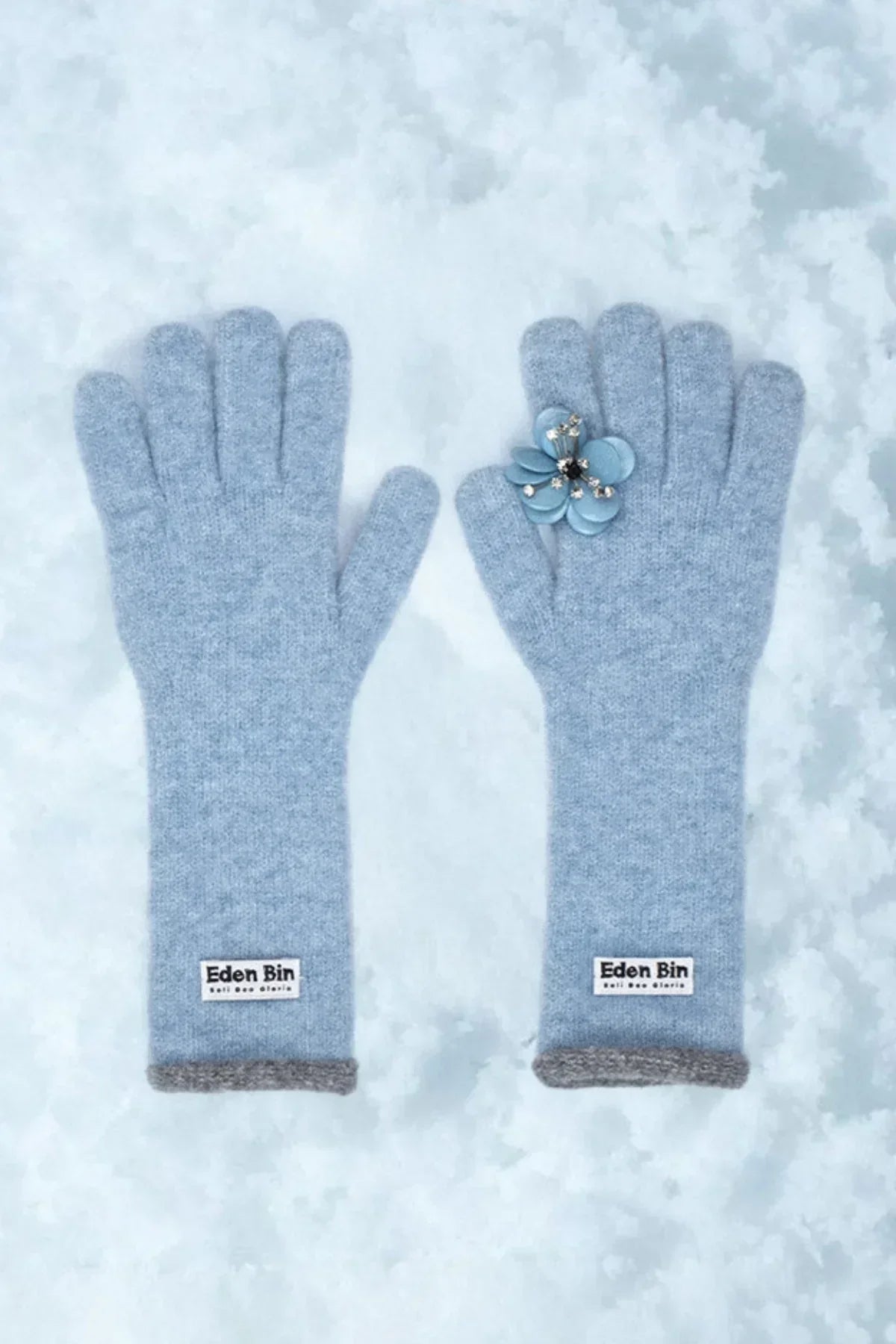 Pair of light blue Eden Bin wool gloves on snow with a blue flower brooch on one glove