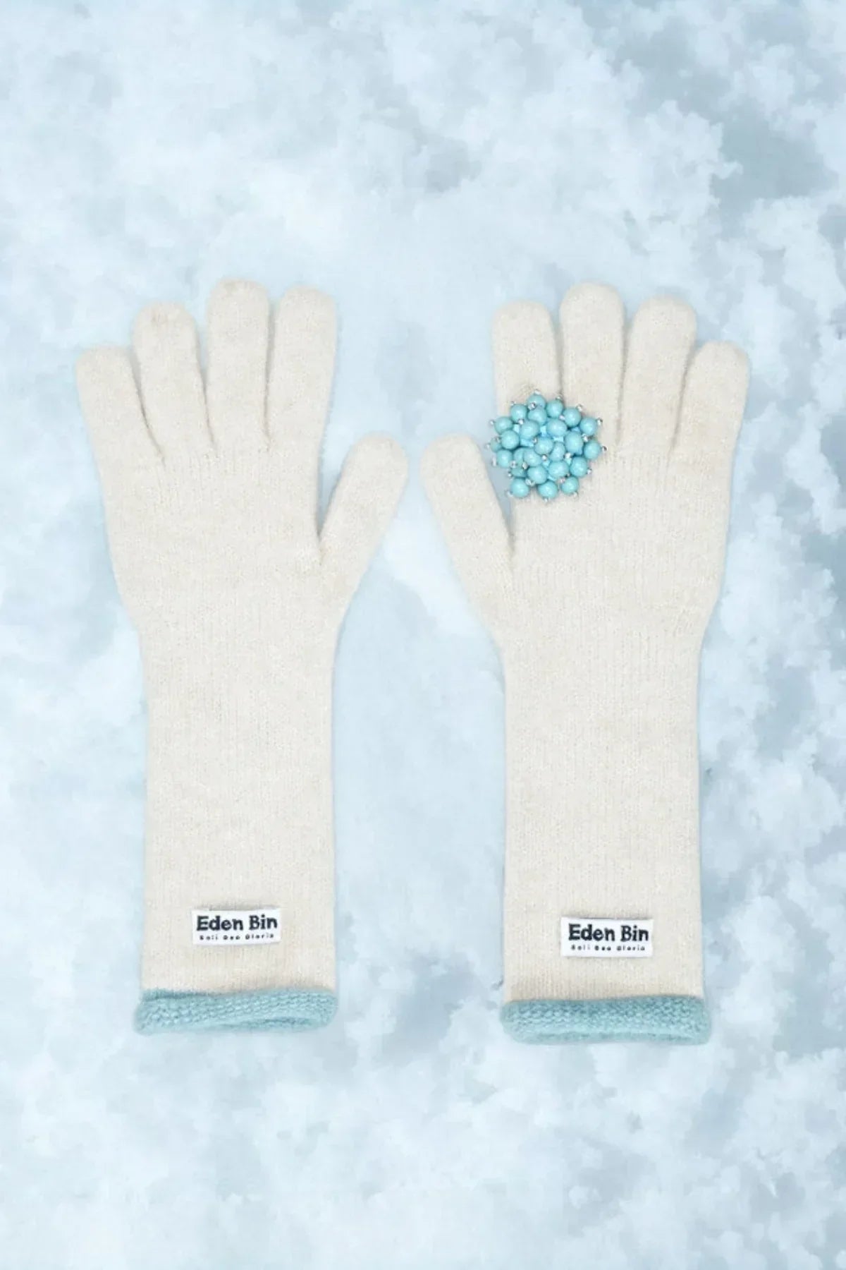 Pair of ivory knit gloves with mint trim and Eden Bin label on light blue textured background, blue bead ring
