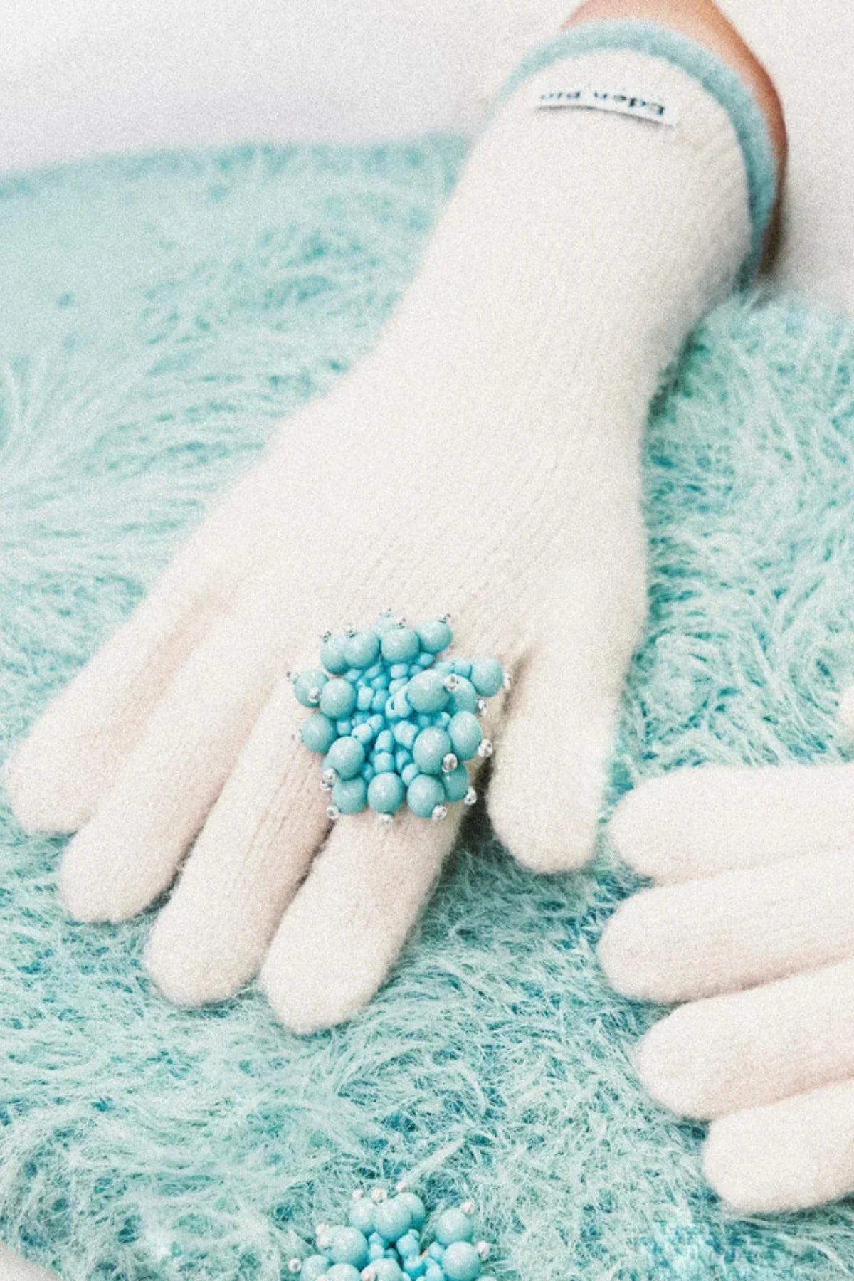 White knitted gloves with a blue beaded flower ring on a textured teal surface