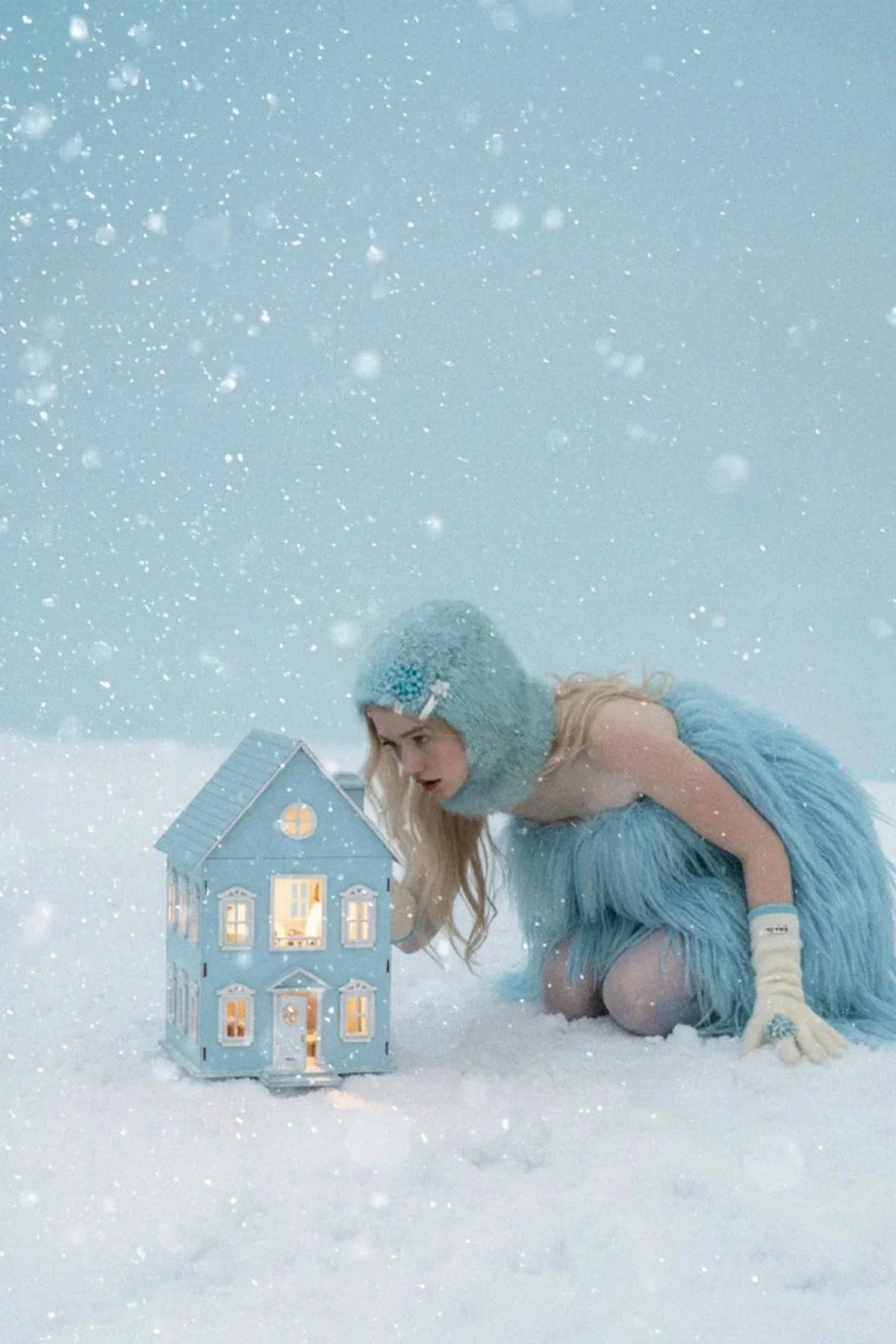 Woman in blue furry outfit and gloves kneeling in snow, looking inside a lit miniature blue house