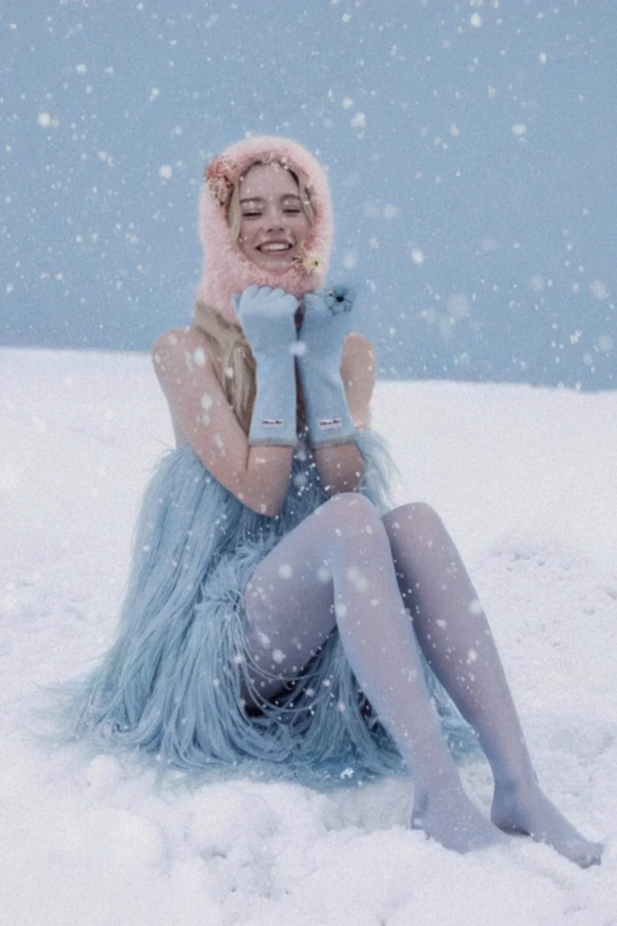 Smiling woman in blue fringed dress, pink furry hood, and blue gloves sitting in snow with snowfall