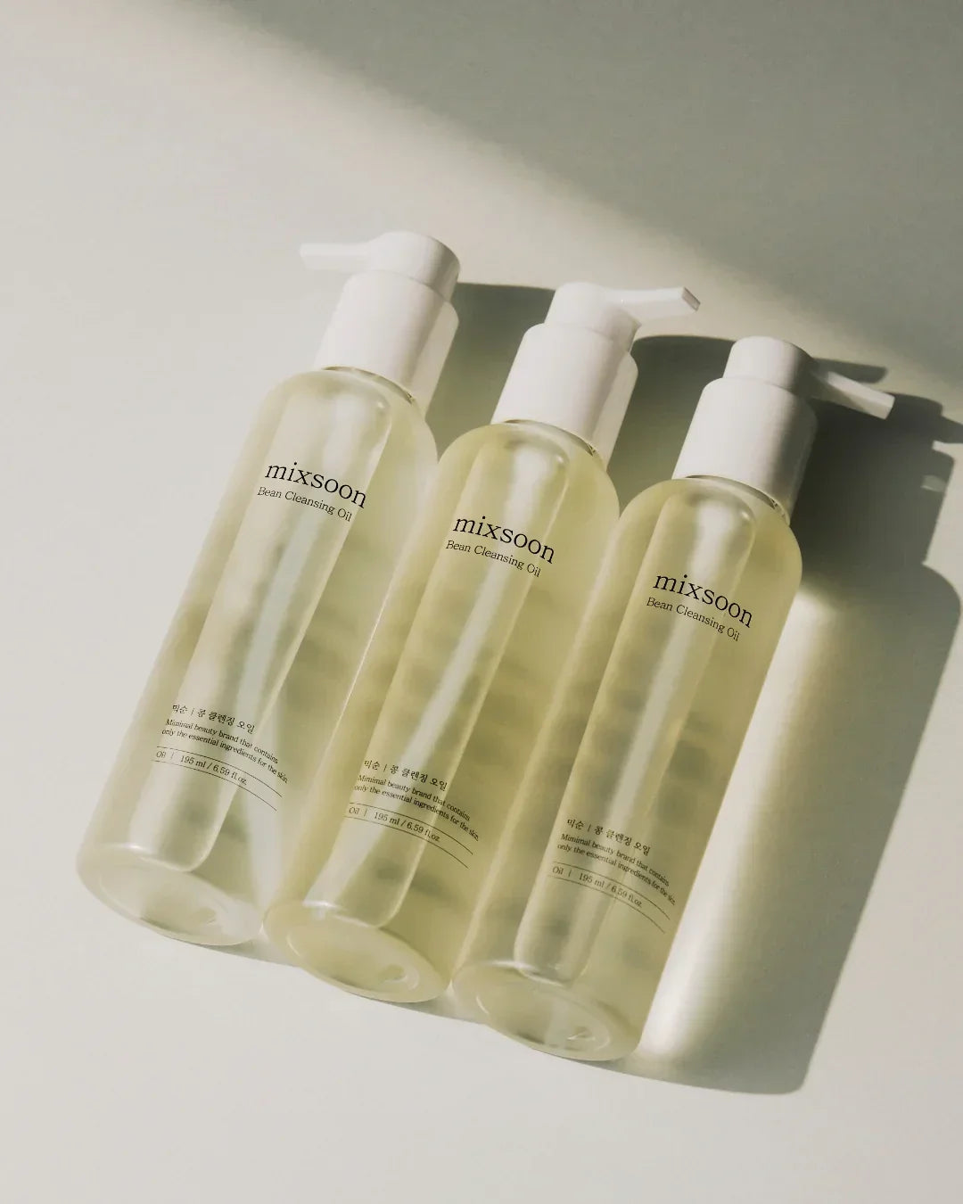 Three transparent bottles of Mixsoon Bean Cleansing Oil with white pump caps on a light surface with shadows