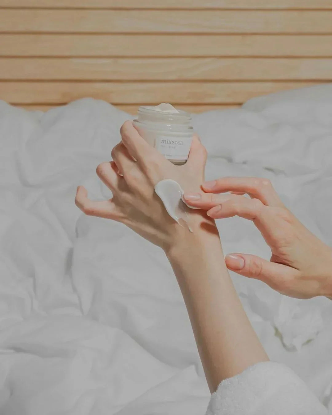 Hands applying Mixsoon bean cream on wrist with soft white bedding and wooden background