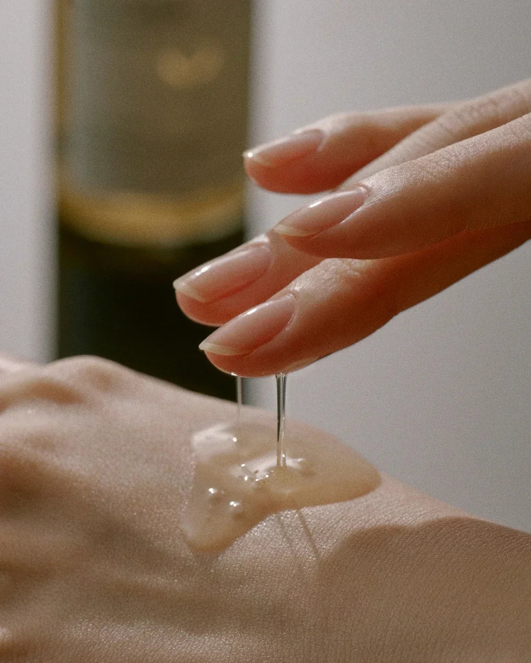 Close-up of fingers applying clear skincare serum on back of hand with blurred bottle in background