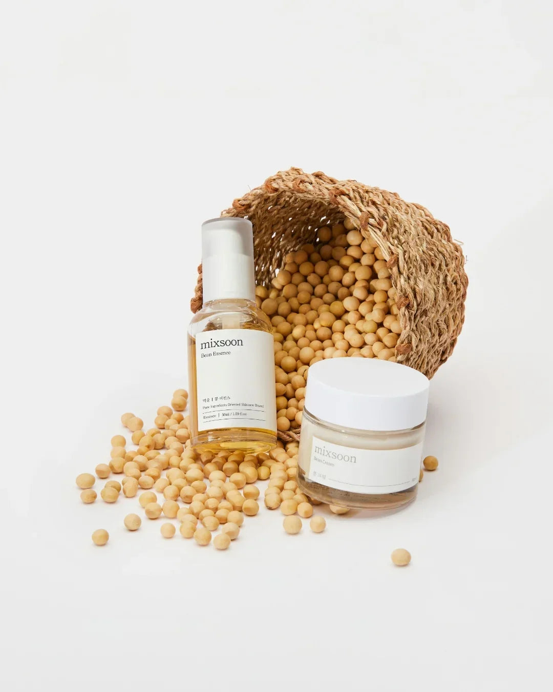 Mixsoon Bean Essence serum and Bean Cream jar with spilled soybeans from woven basket on white background