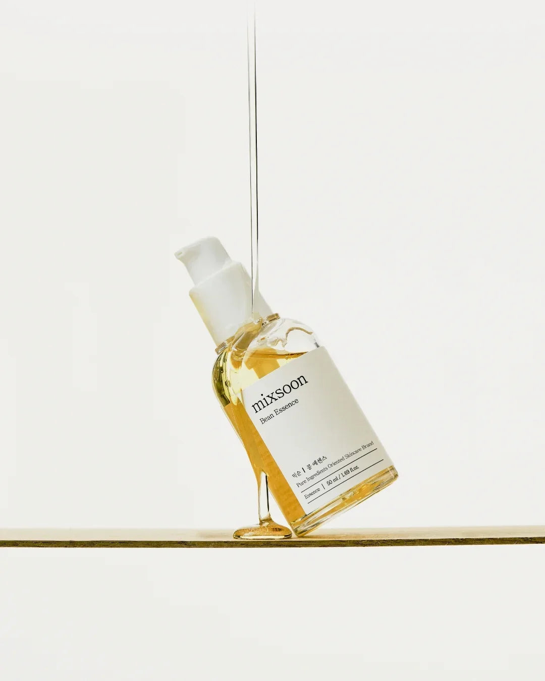 Mixsoon Bean Essence skincare bottle pouring clear serum on wooden surface in minimal white background