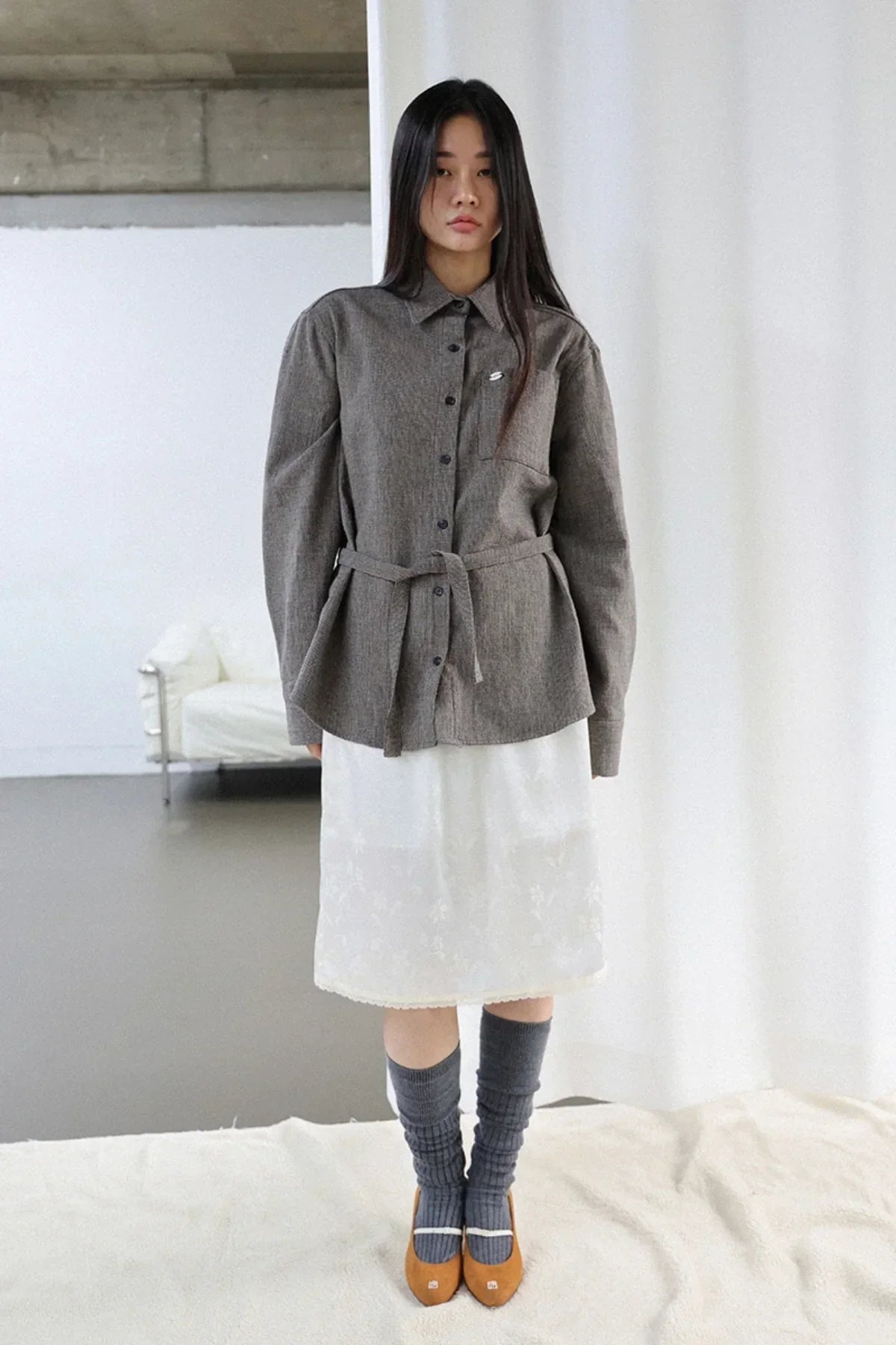Model wearing brown belted button-up shirt, white skirt, gray knee-high socks, and brown shoes in minimal studio