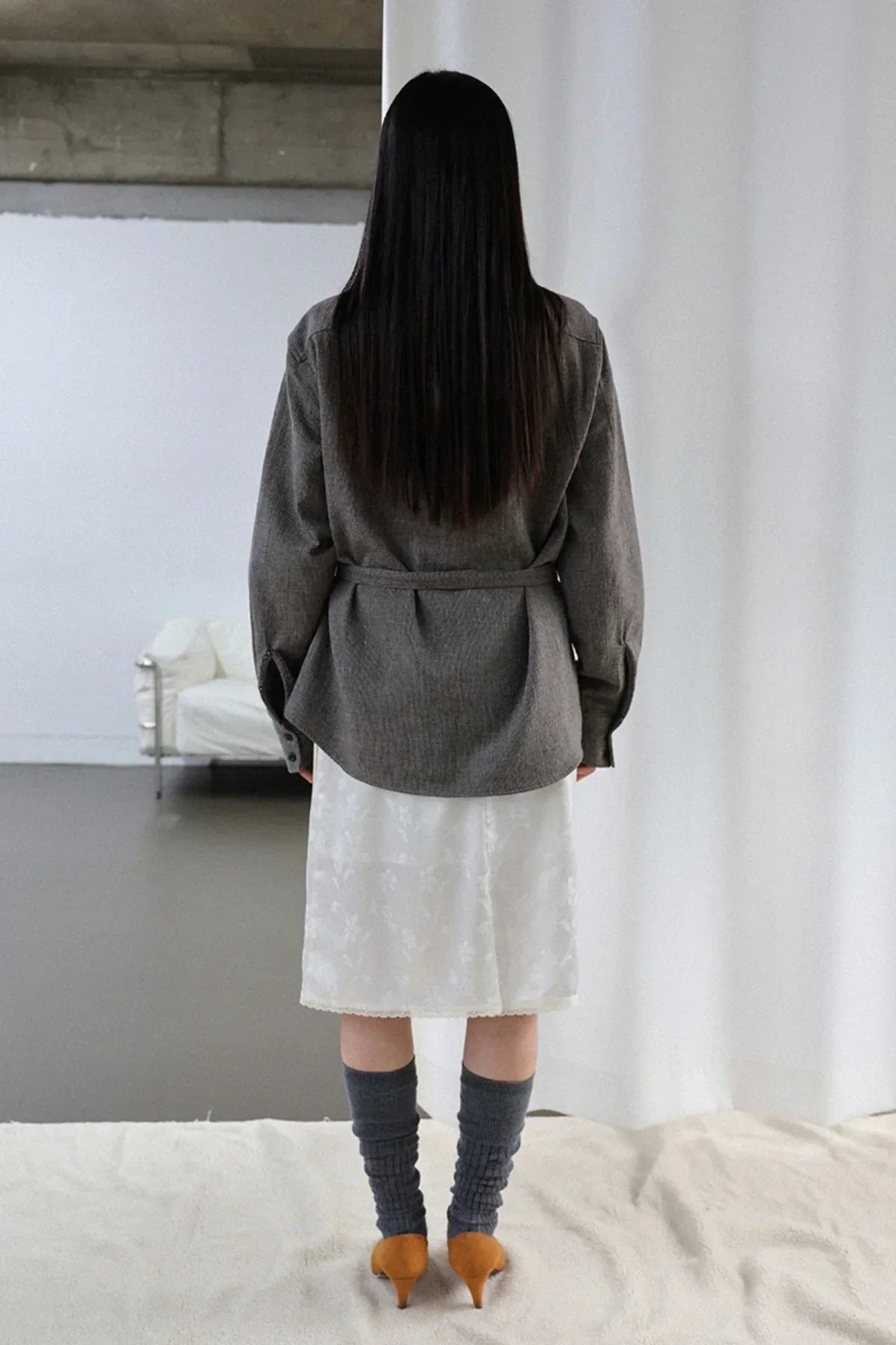 Woman with long black hair wearing belted gray shirt, white skirt, gray socks, and orange heels indoors