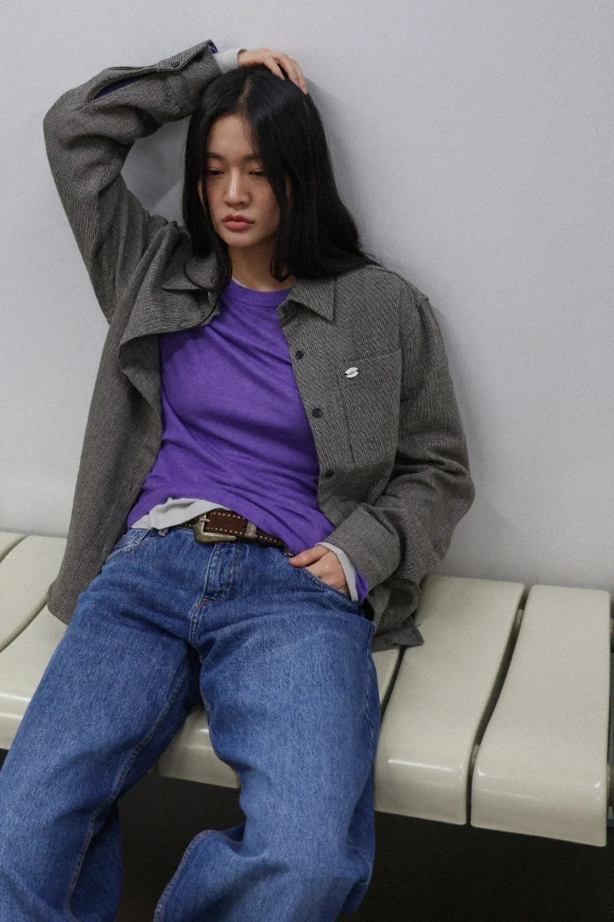 Woman in purple shirt, gray jacket, and blue jeans sitting on white bench against gray wall