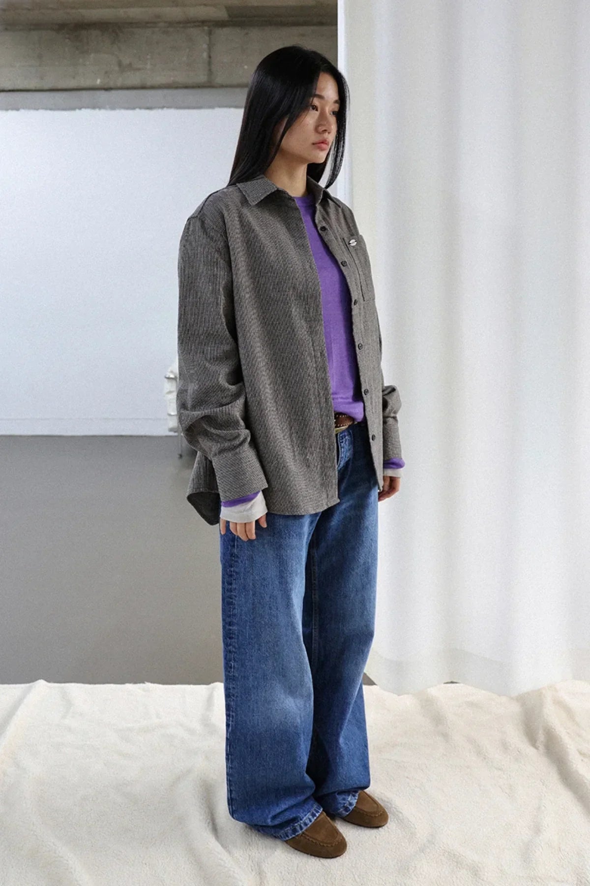Young woman in purple shirt, gray oversized jacket, wide blue jeans, and brown shoes in minimal indoor setting