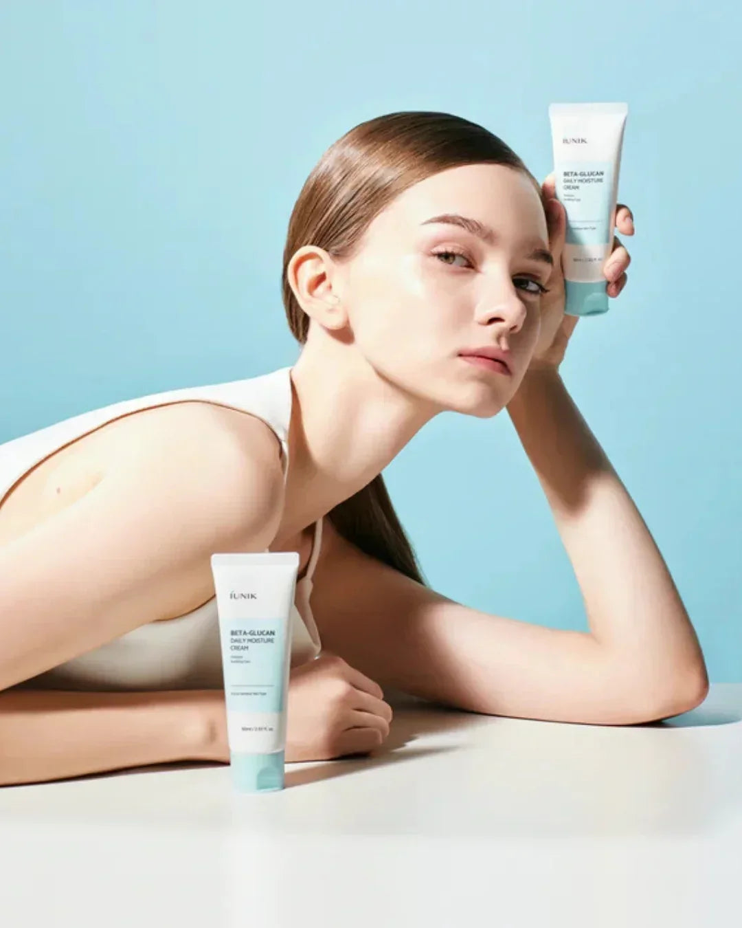 Model with sleek hair posing with iUNIK Beta-Glucan Daily Moisture Cream against light blue background