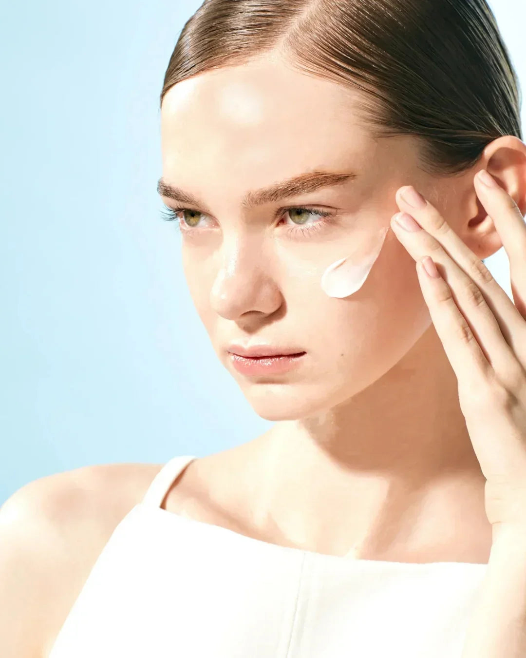 Young woman applying cream on clean skin with blue background, skincare moisturizing routine