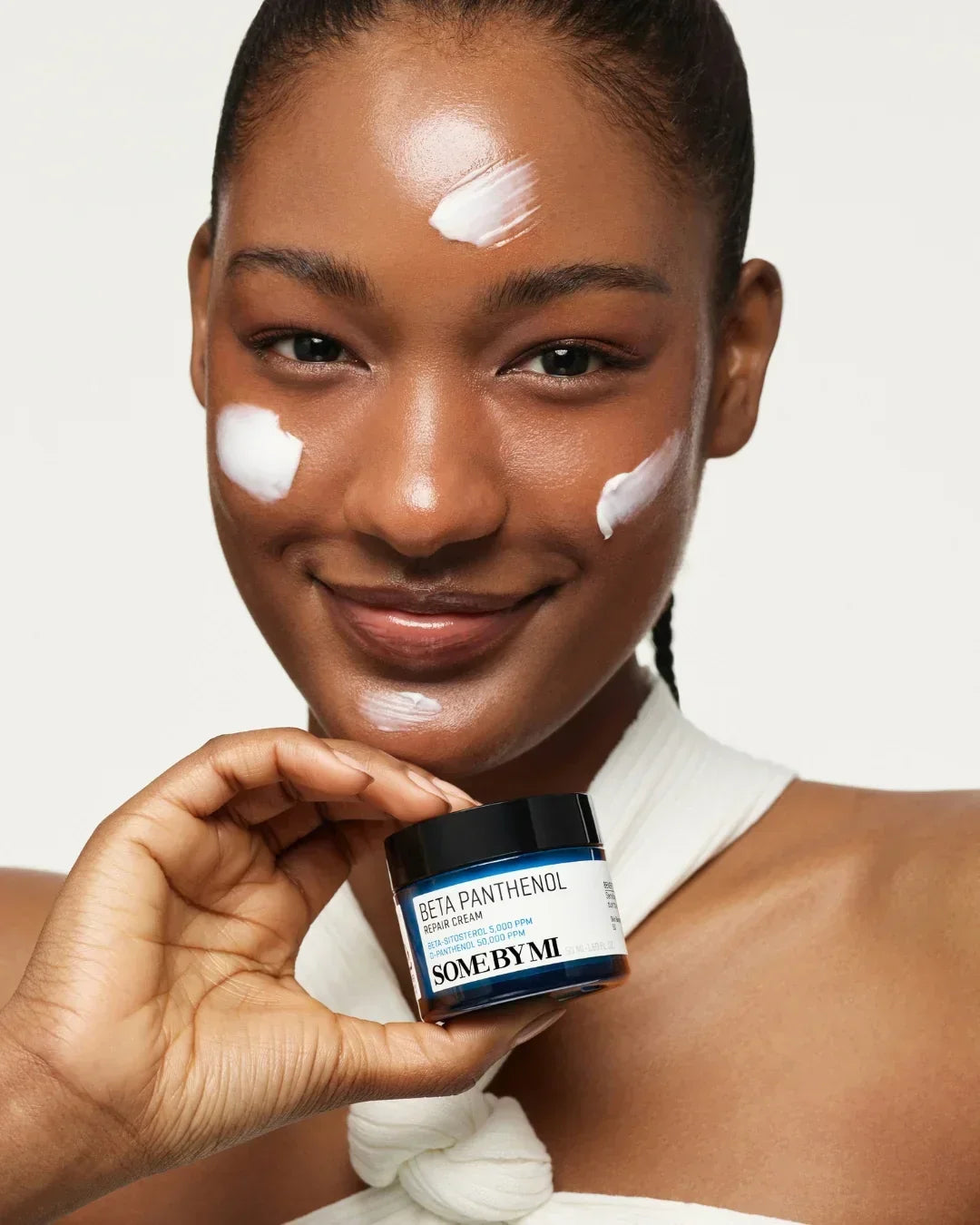 Smiling woman with glowing skin holding Some By Mi Beta Panthenol Repair Cream with cream dots on her face