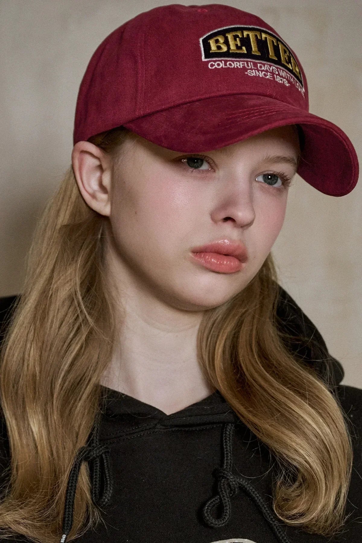 Close-up of young woman wearing a burgundy baseball cap with 'BETTER' embroidered and a black hoodie