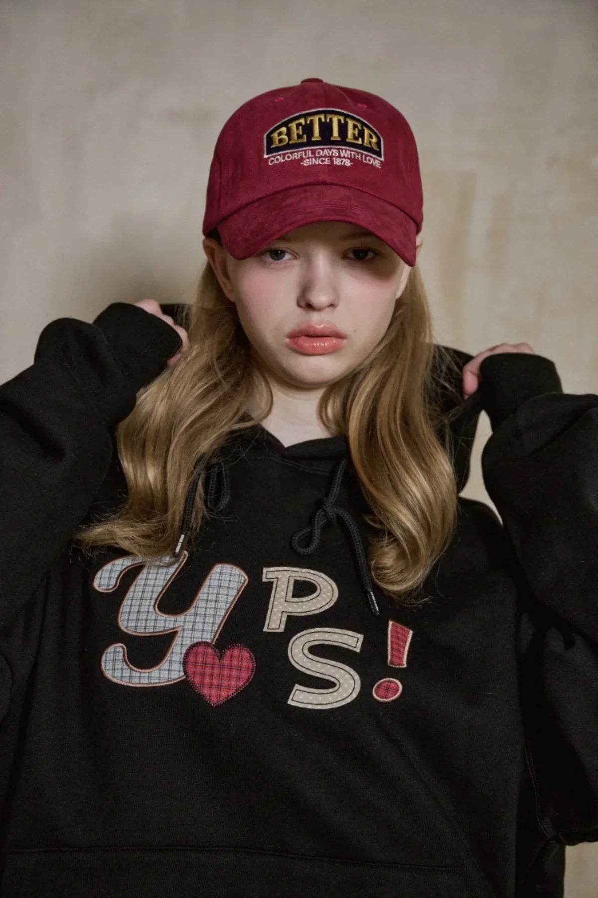 Young woman wearing a burgundy baseball cap with 'BETTER' and black hoodie with colorful 'YUPS!' print