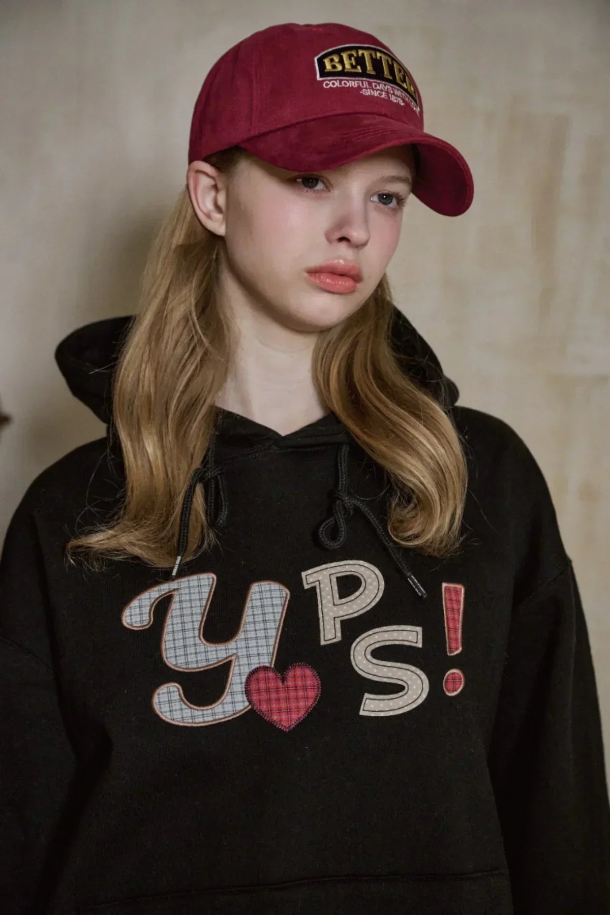 Young woman wearing a burgundy Better Ballcap and a black hoodie with 'YPS!' text and heart design