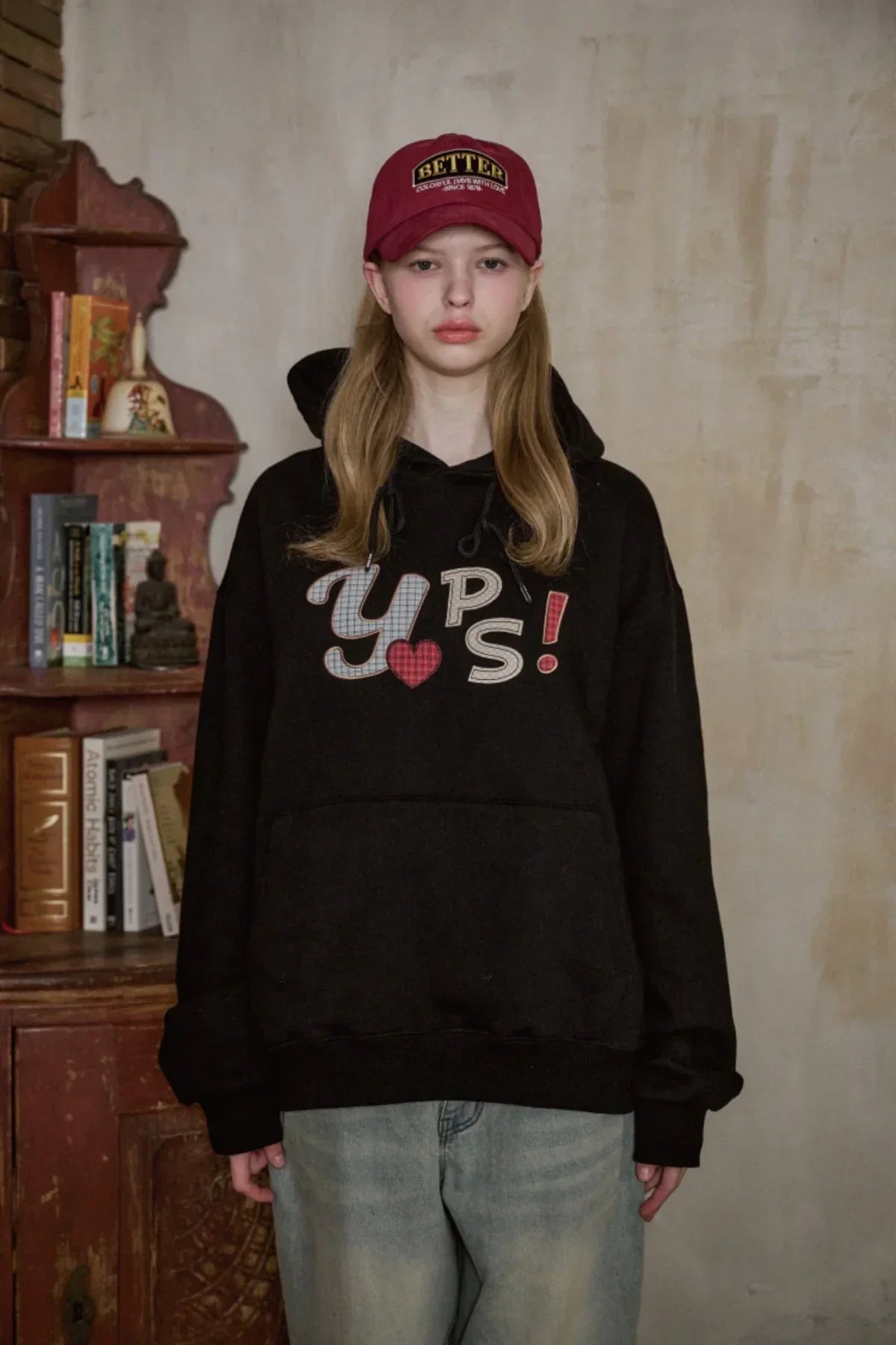 Young woman wearing a burgundy Better baseball cap and black YPS! hoodie in a rustic indoor setting