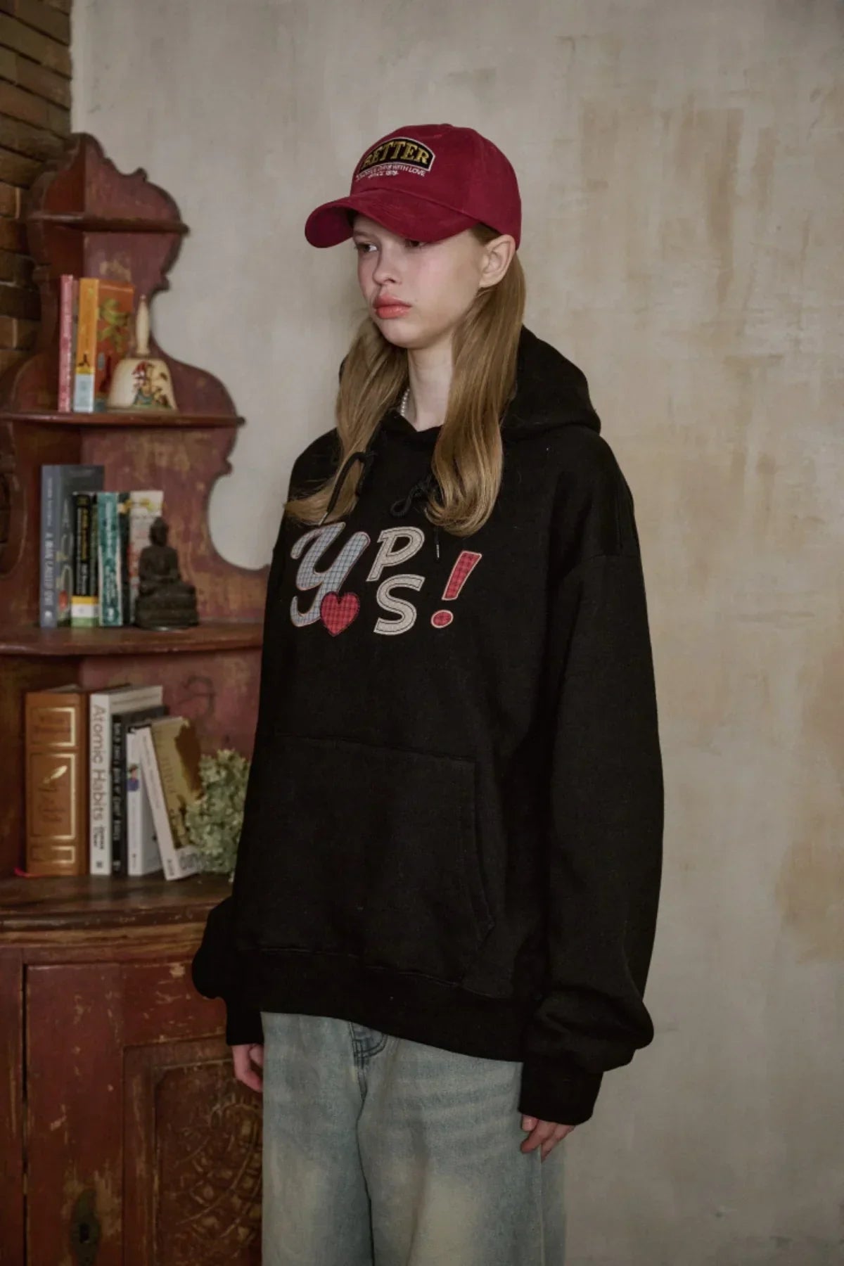 Young woman wearing burgundy Better ballcap and black hoodie with graphic text, standing indoors near vintage wooden shelf