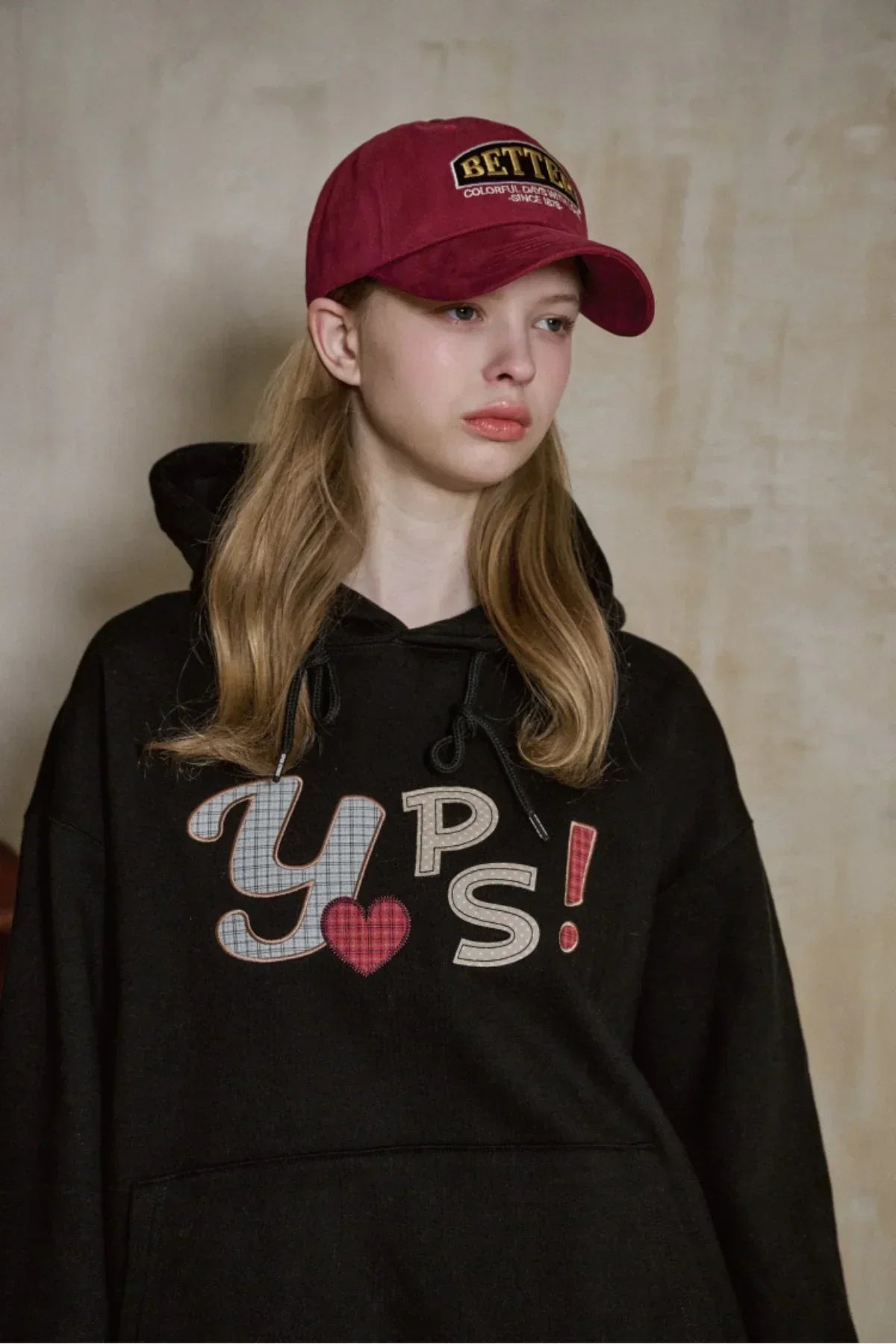Young woman wearing a burgundy Better ballcap and black hoodie with YPS! logo in a neutral studio