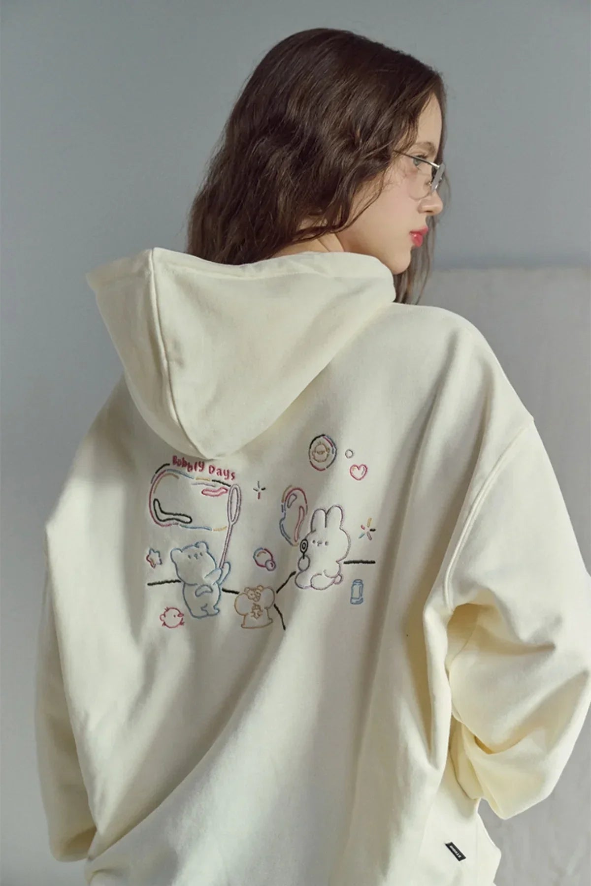Woman wearing cream hoodie with colorful embroidered cartoon characters on back, posing indoors