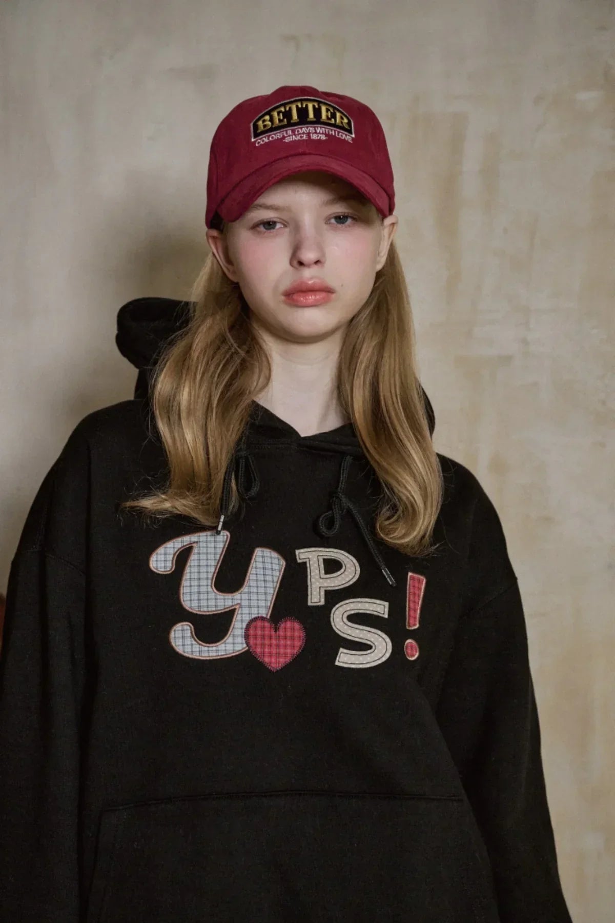 Young woman wearing burgundy BETTER ballcap and black hoodie with colorful YPS! lettering on beige background