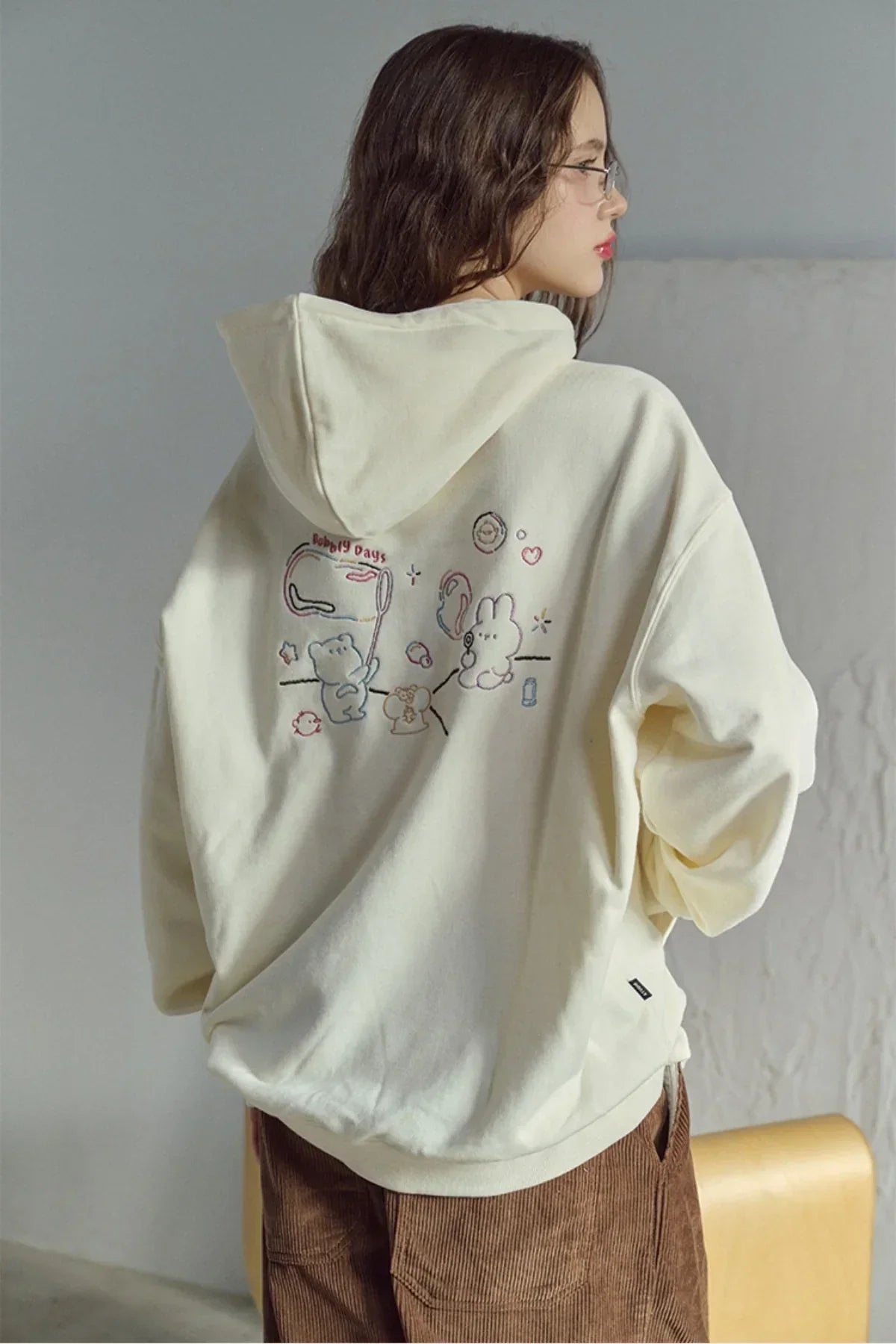 Woman wearing cream hoodie with cute embroidered cartoon bears and bunny design on back indoors
