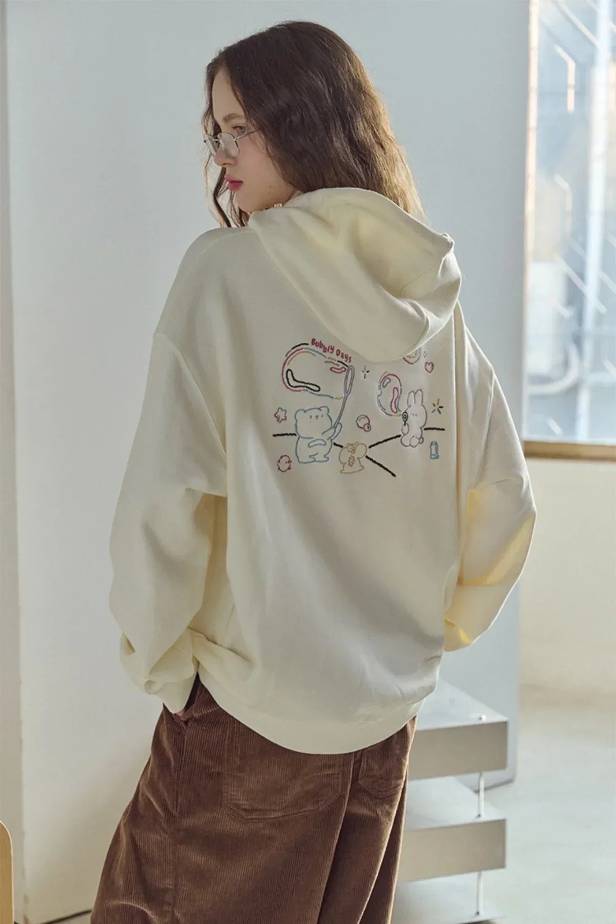 Woman wearing cream hoodie with cute cartoon bear and bunny graphic on back, brown corduroy pants indoors