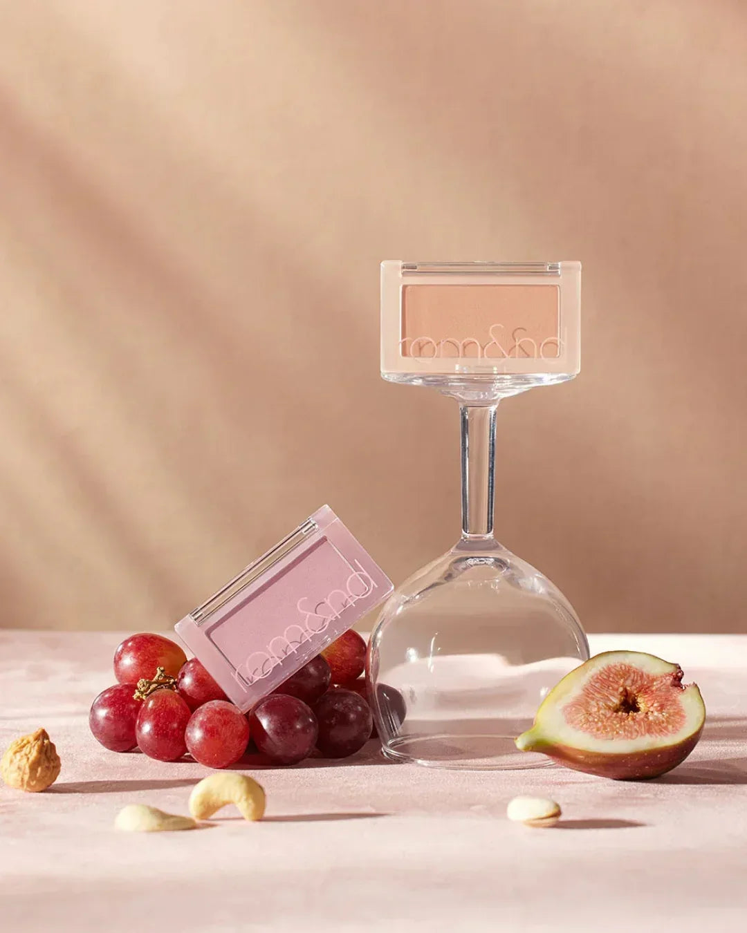 rom&nd blush compacts on grapes and upside-down wine glass with fig and nuts on pink surface