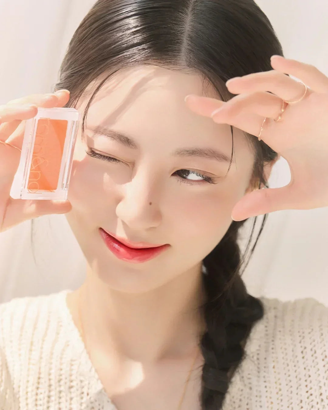 Young woman with braided hair holding orange cream blush compact, smiling with sunlit face