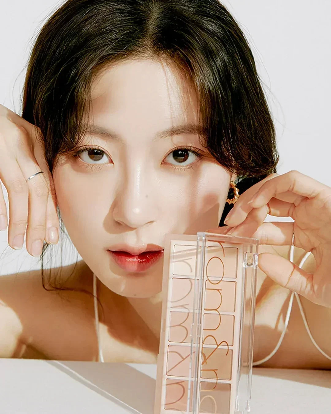 Close-up of woman holding a nude eyeshadow palette with clear lid against white background