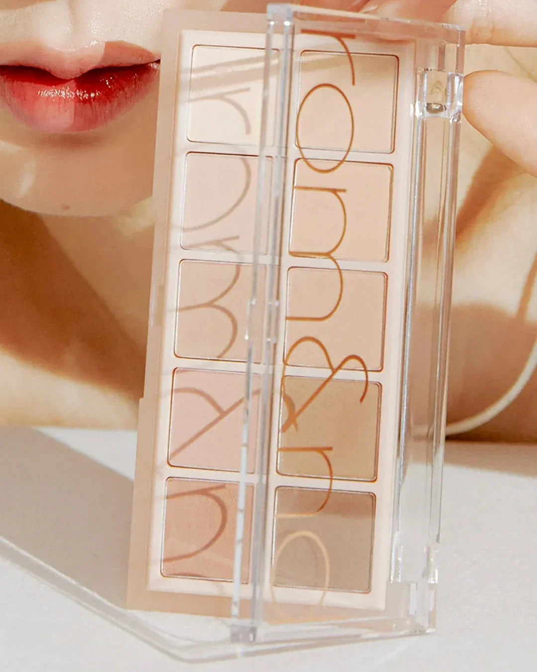 Close-up of a face with red lips holding an eyeshadow palette with neutral shades