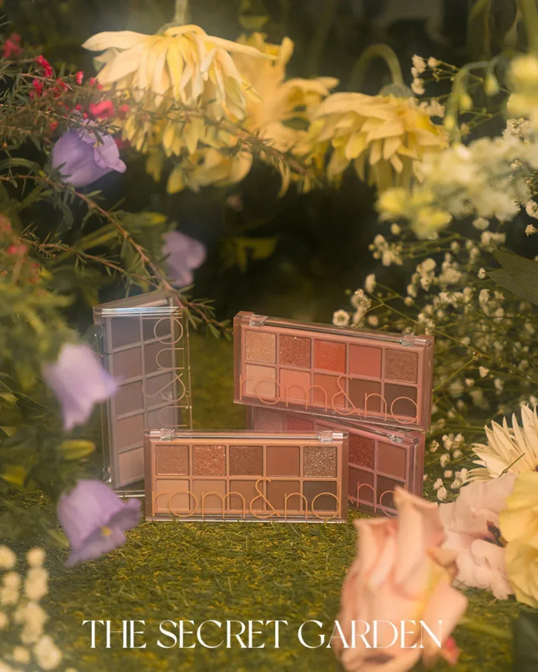 Rom&nd eyeshadow palettes displayed on grass surrounded by colorful flowers in a secret garden setting