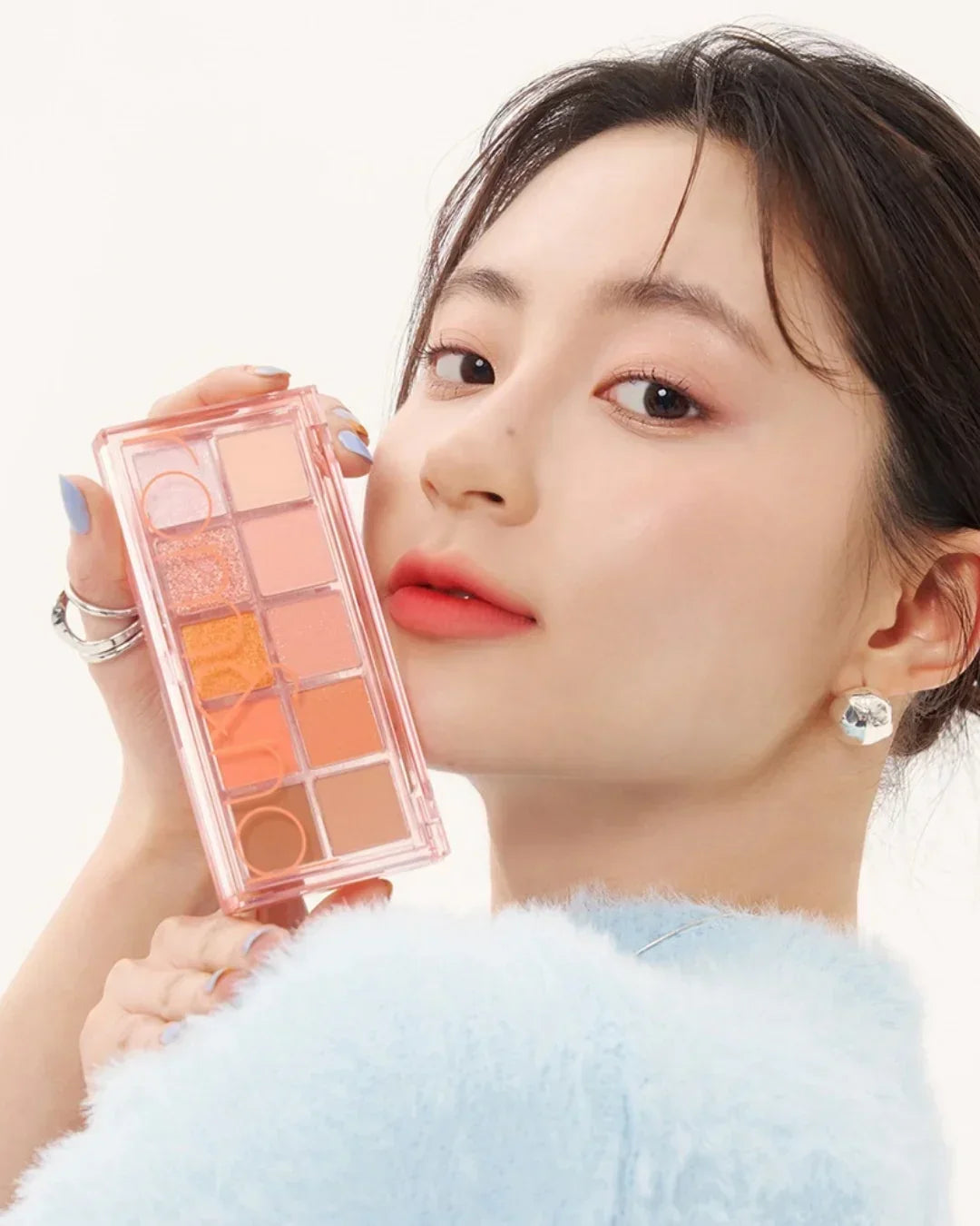 Close-up of woman in blue fuzzy sweater holding peach-toned eyeshadow palette near face