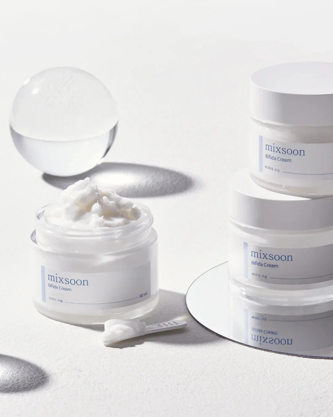 Mixsoon Bifida Cream jars with white cream and spatula on white surface with glass sphere and mirror
