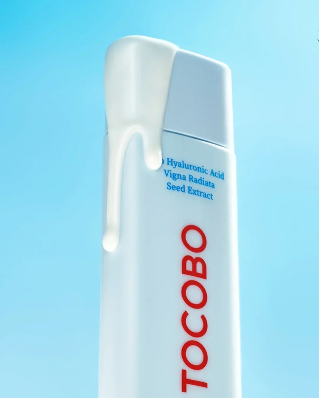 White TOCOBO skincare bottle with dripping cream against blue background, featuring hyaluronic acid and seed extract