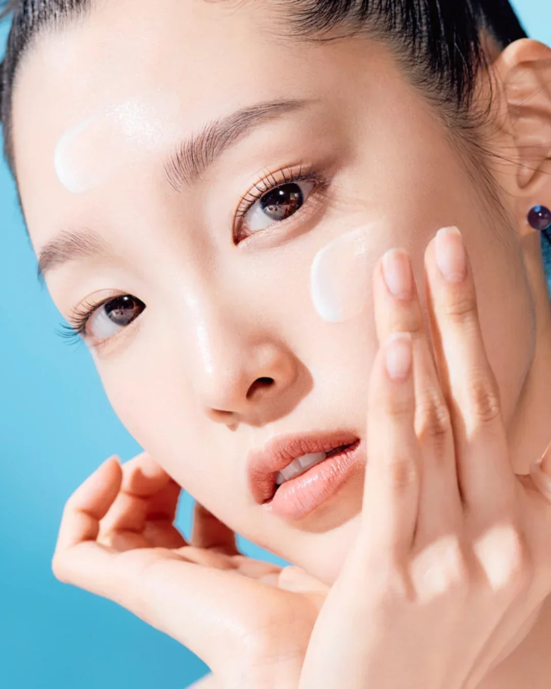 Close-up of woman applying white sunscreen cream on cheek and forehead with blue background