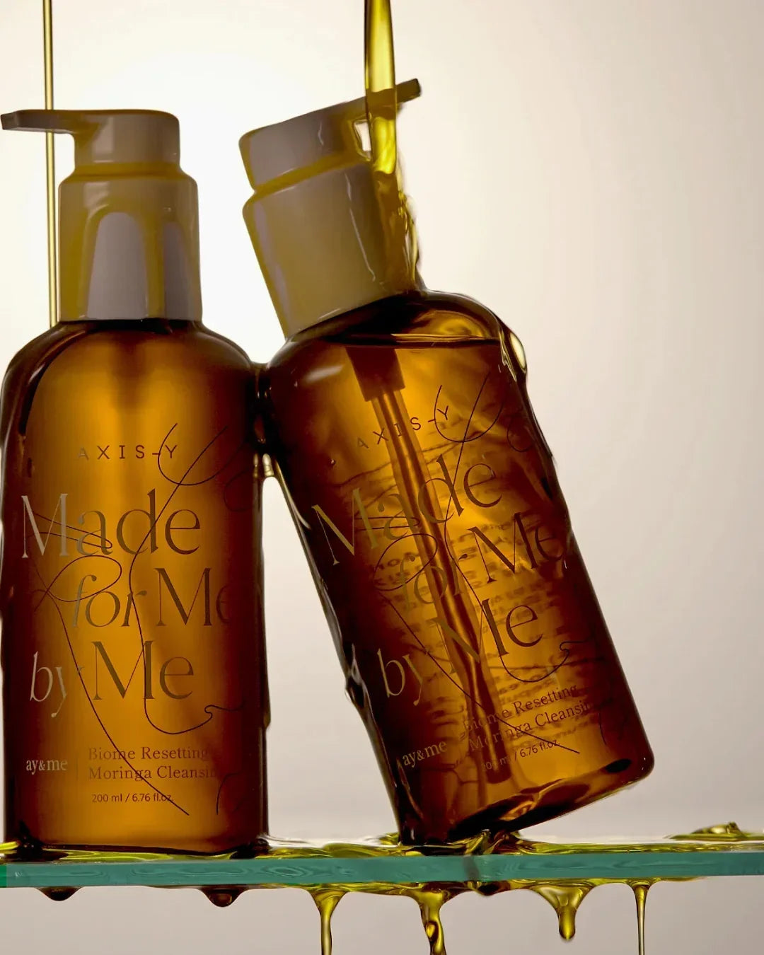 Two amber pump bottles of AXIS-Y Made for Me Biome Resetting Moringa Cleansing Oil with oil dripping down