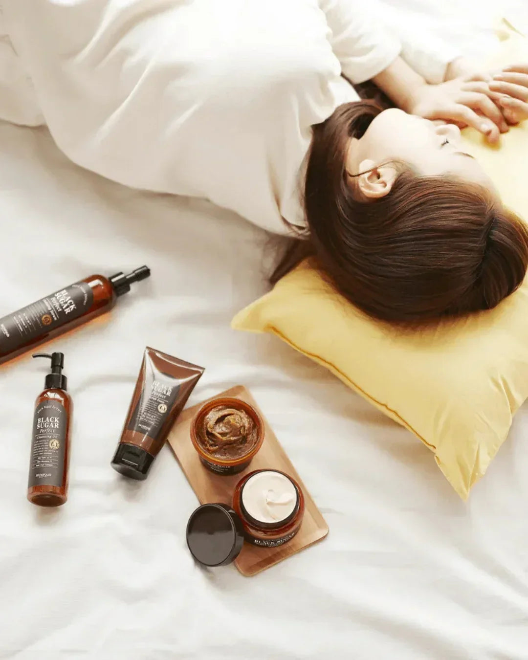 Woman lying on white bed with yellow pillow surrounded by black sugar skincare products