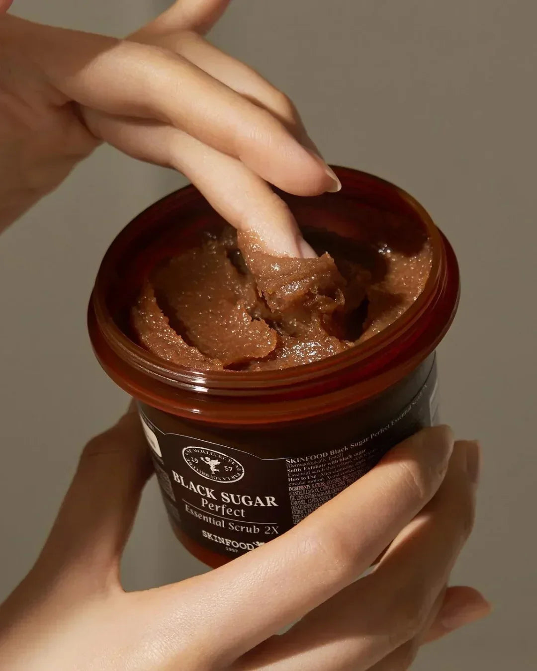 Hands scooping SKINFOOD Black Sugar Perfect Essential Scrub 2X from brown jar under soft lighting