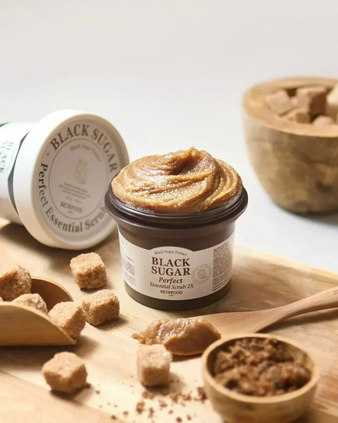 Black Sugar Perfect Essential Scrub 2X open jar with scrub and brown sugar cubes on wooden surface