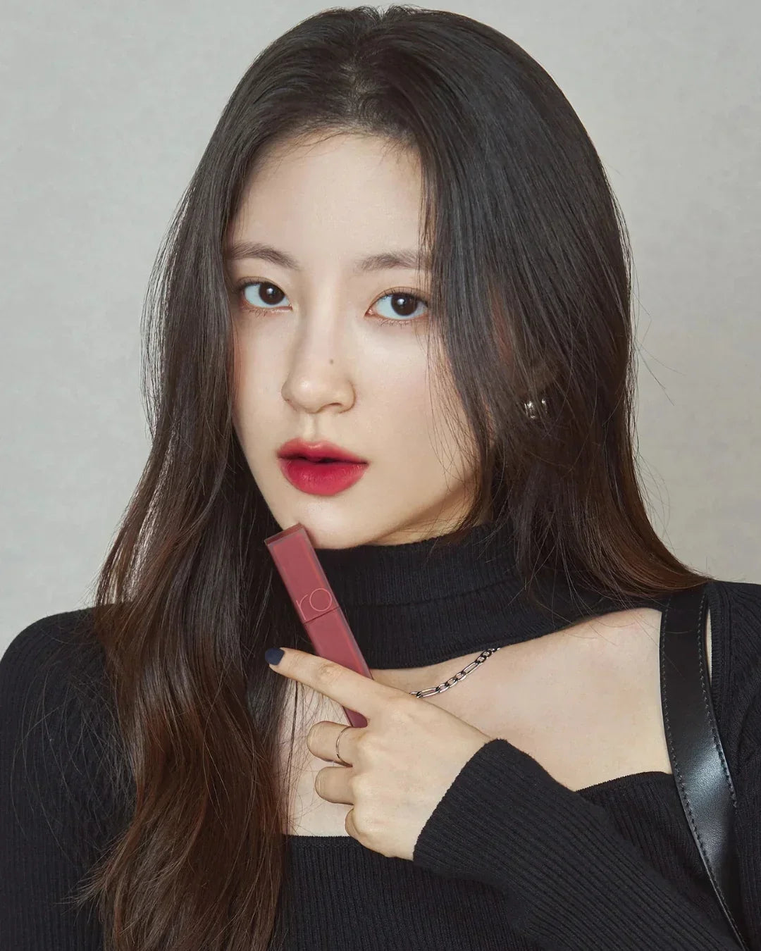 Young woman with long dark hair wearing black holding mauve lipstick close to her face
