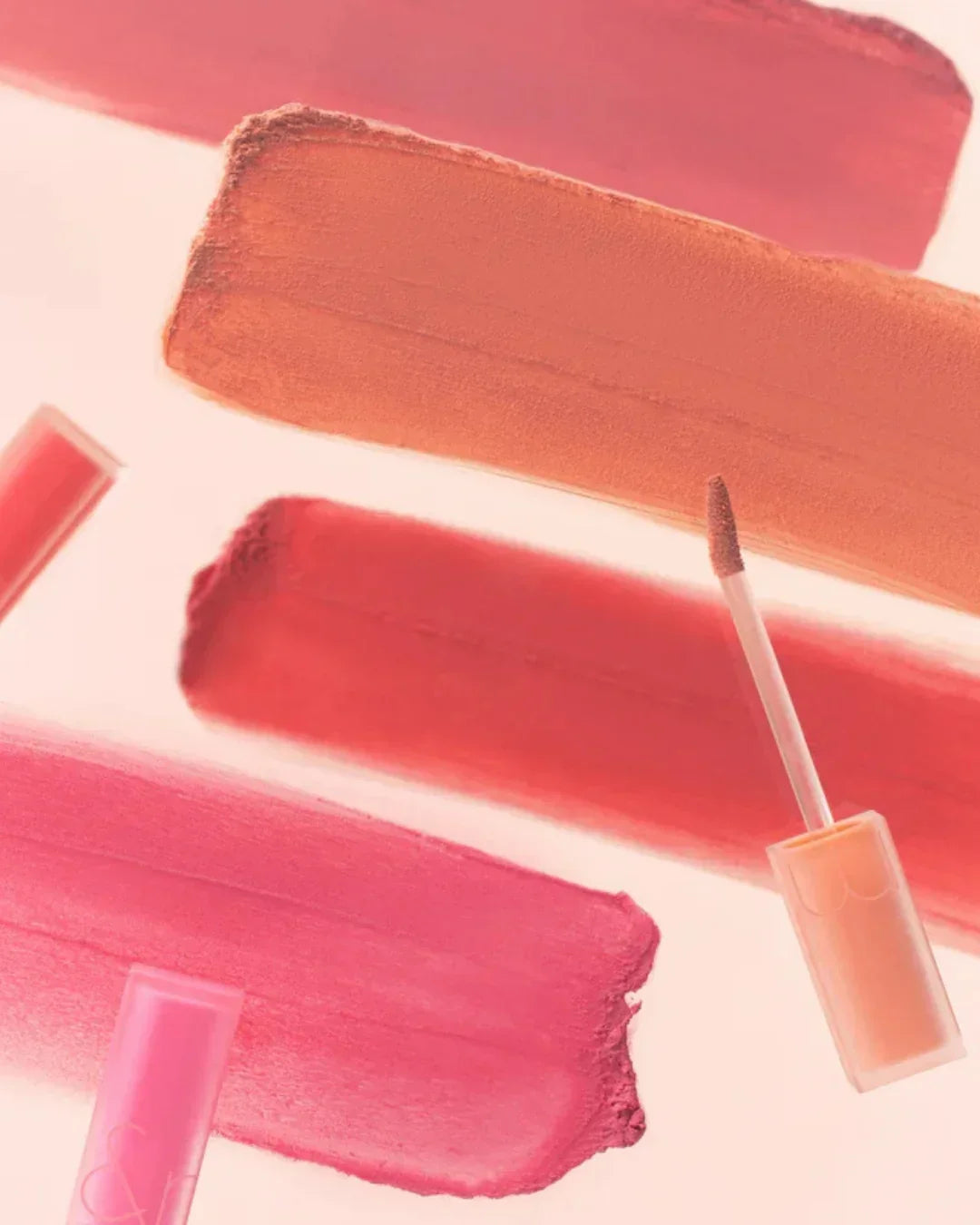 Close-up of coral, pink, and red lip gloss swatches with applicators on light background