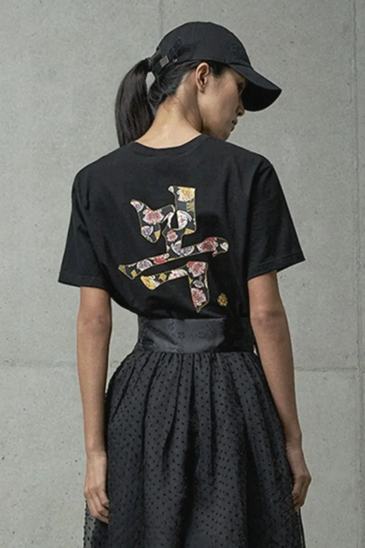 Woman wearing black t-shirt with floral patterned Asian character print on back, paired with black skirt and cap