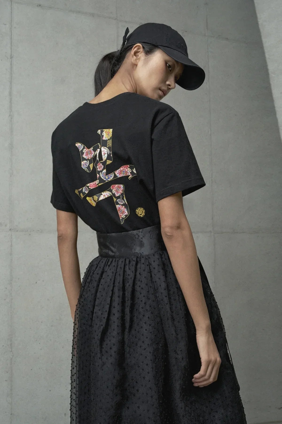 Woman wearing black t-shirt with colorful floral Asian script print on back, black cap, and textured skirt